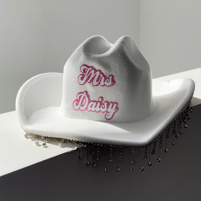 cow girl hat with rhinestone tassles hanging along the brim. surname written in cow boy font with pink and white colourong. hat is white